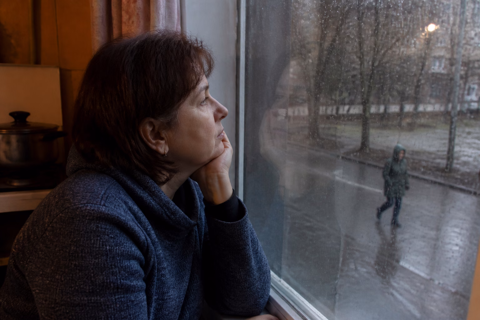 Жінка замислено дивиться крізь запітніле вікно, надворі дощ, розмитий силует перехожого